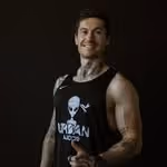 Smiling tattooed man with muscular arms wearing a black Urban legend tank top against dark background.