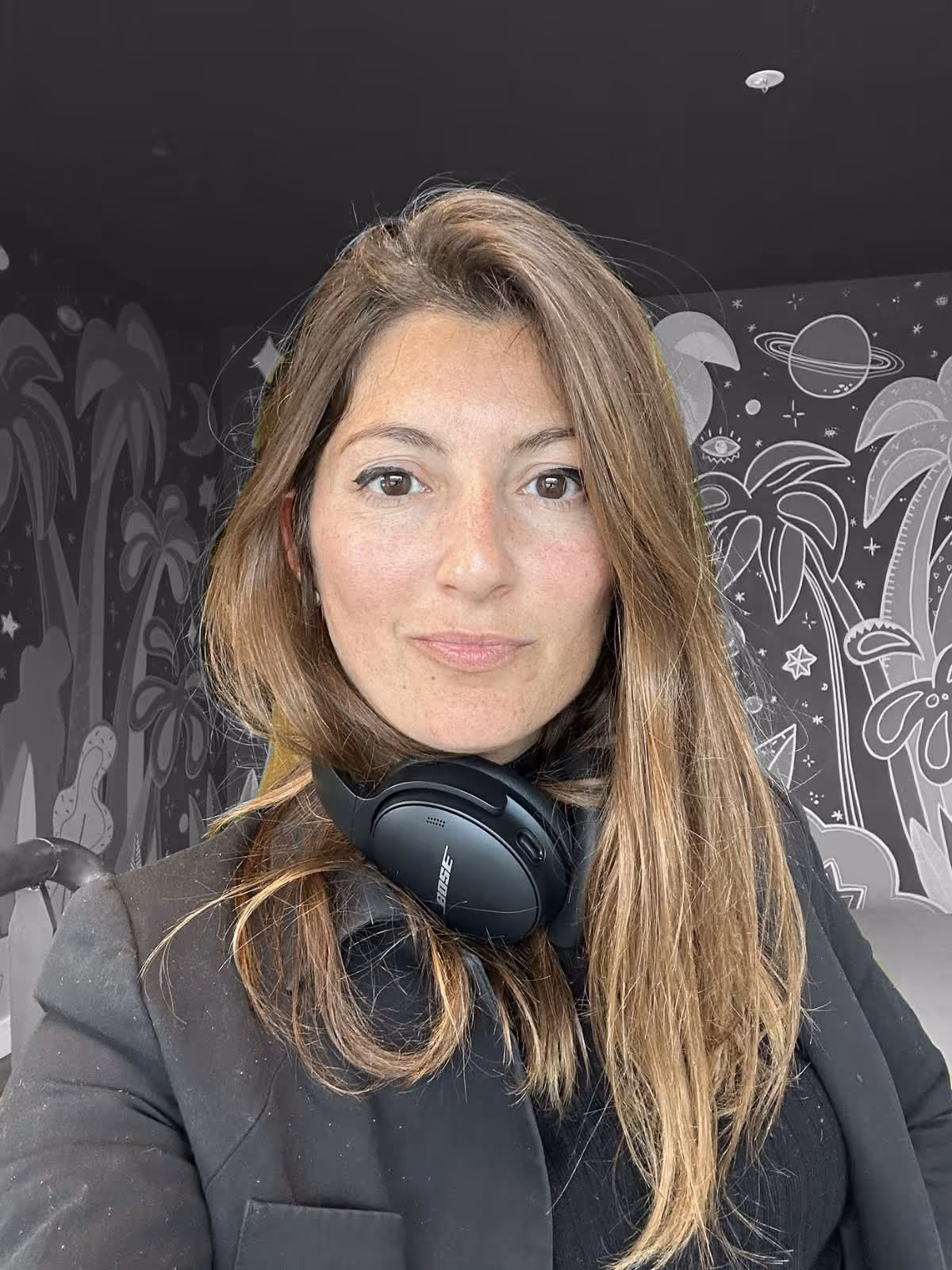 Woman with long hair wearing black blazer and Bose headphones around her neck, with illustrated wall behind.