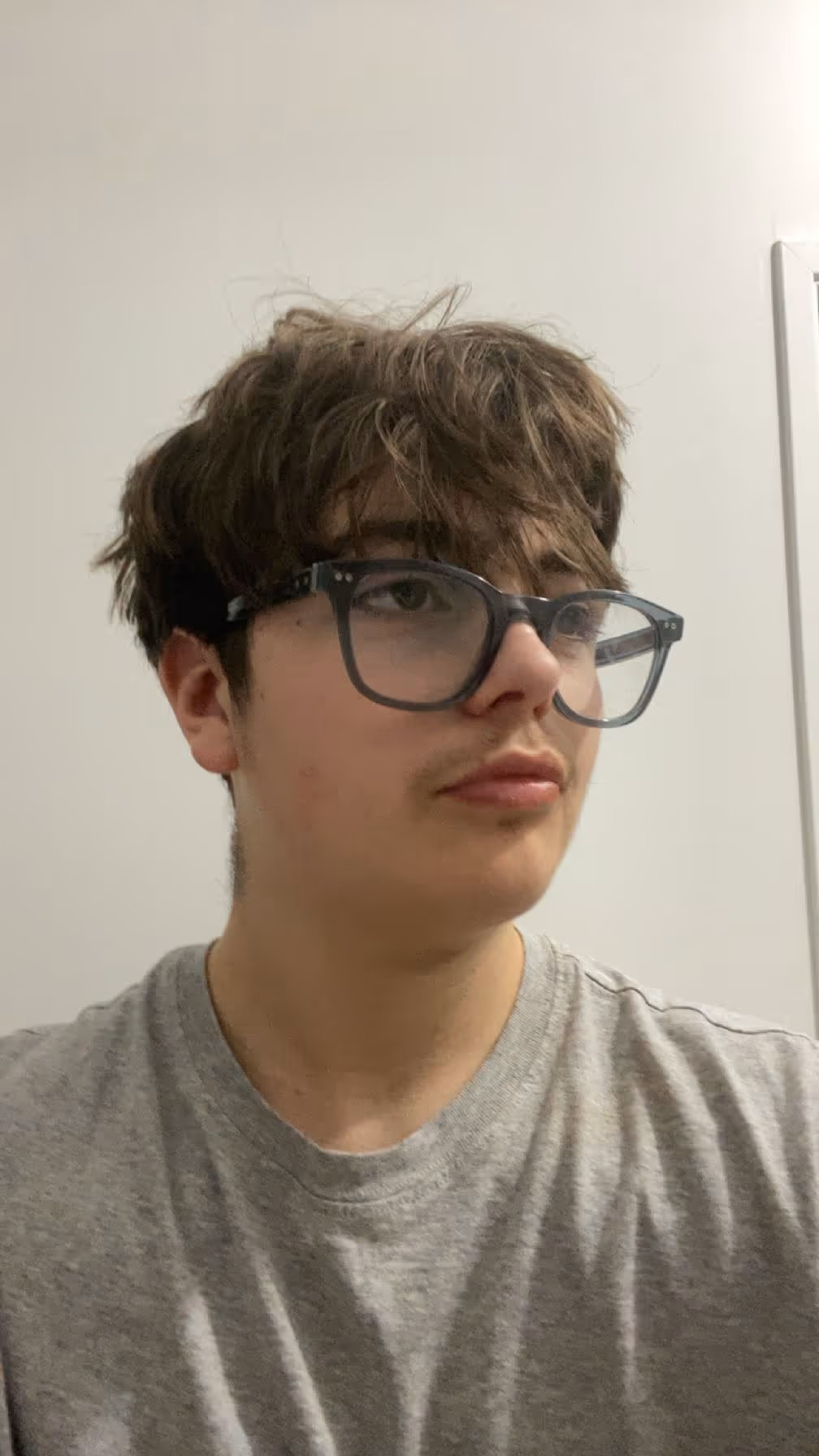 Young person with tousled brown hair and glasses wearing a gray shirt looking to the side indoors.
